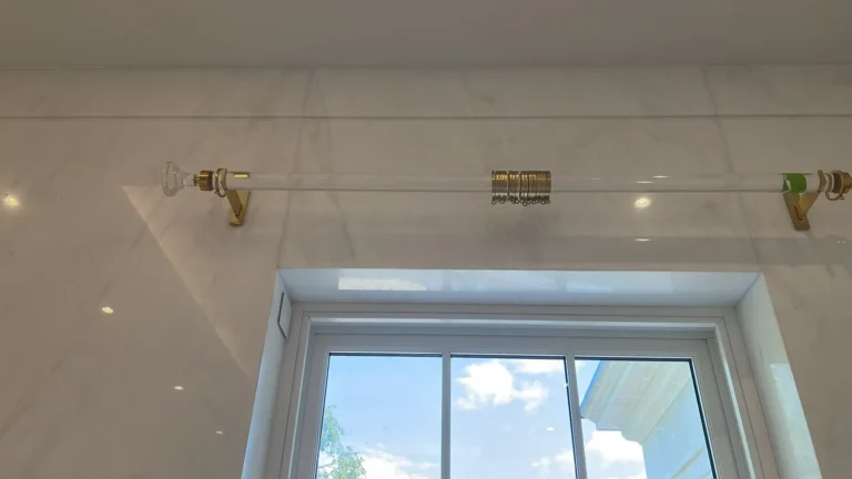 Clear acrylic curtain pole with brass rings above a window in a marble-clad bathroom