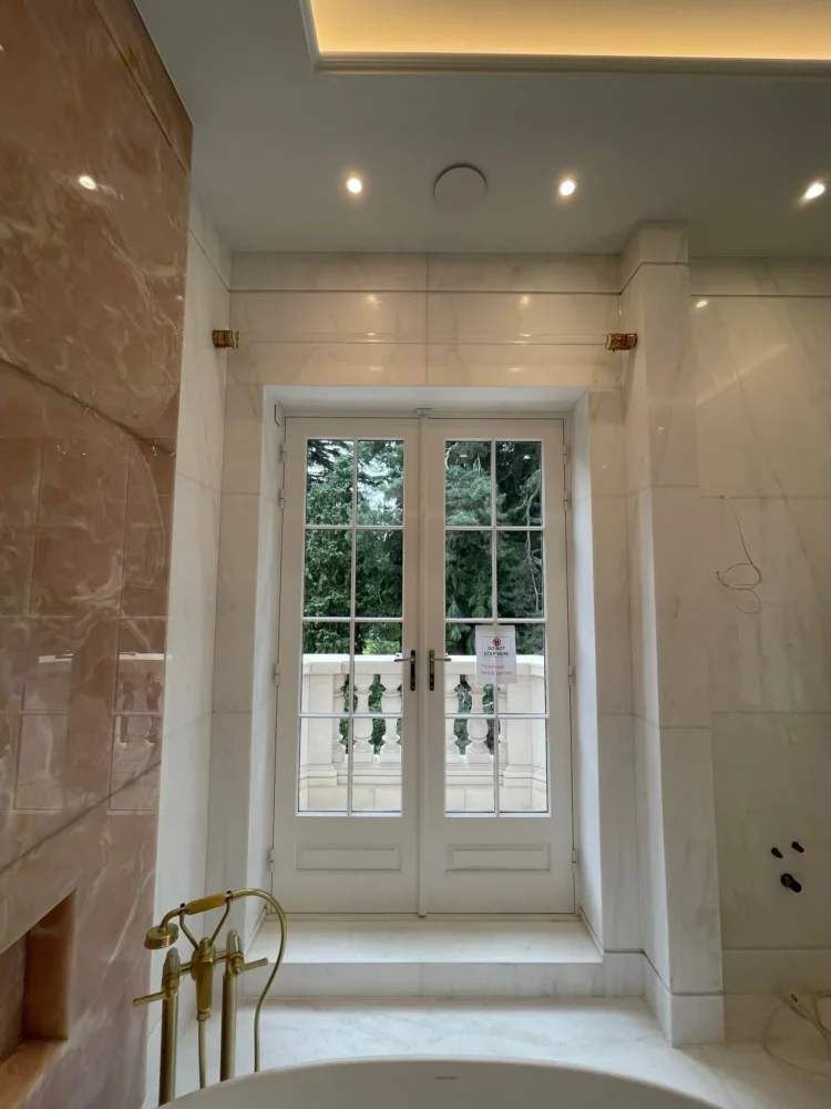 Brass pole brackets fitted above French doors in a marble bathroom with a freestanding bath filler