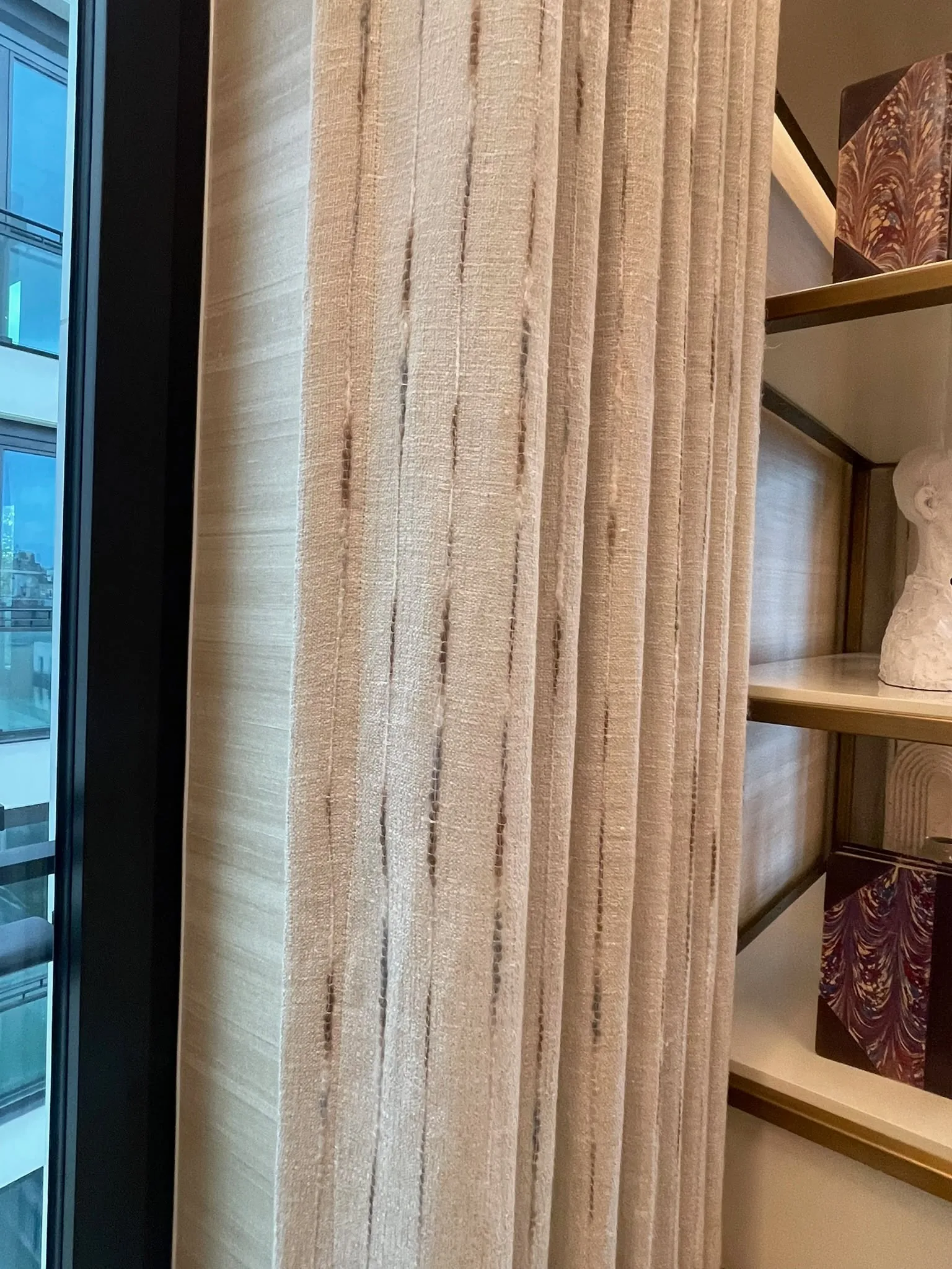 Striped textured curtain hanging in tall folds beside a full-height window and display shelves