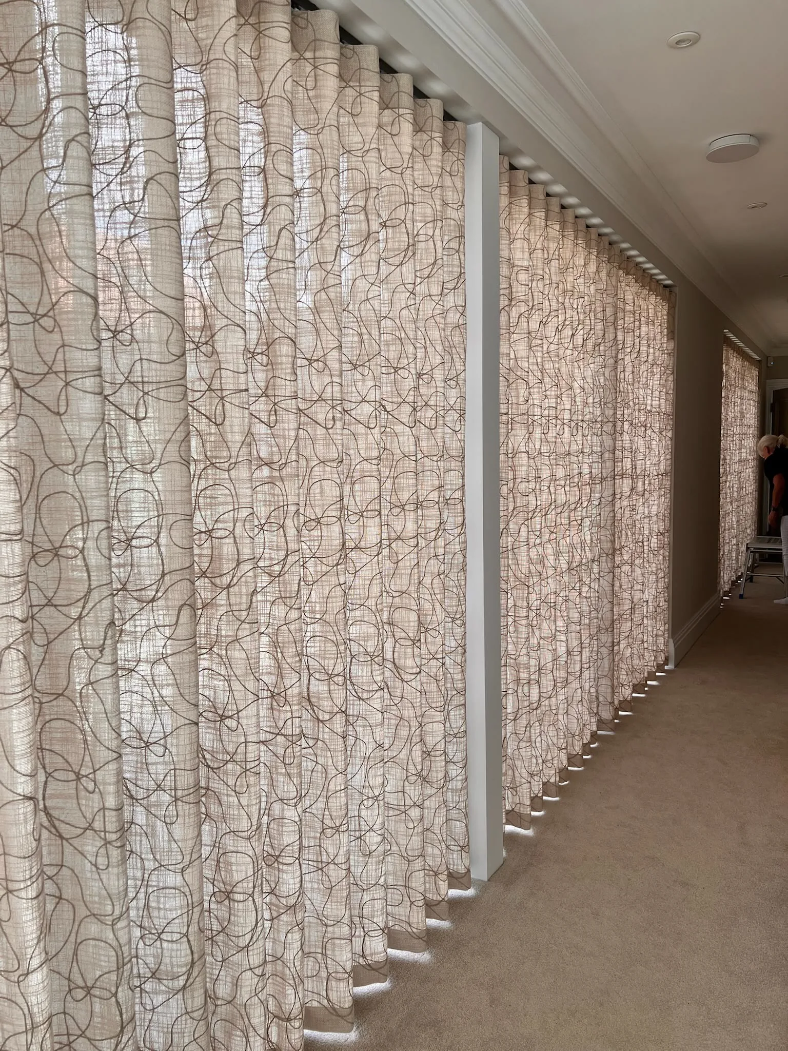 Long run of patterned sheer wave curtains on a ceiling track along a corridor of windows