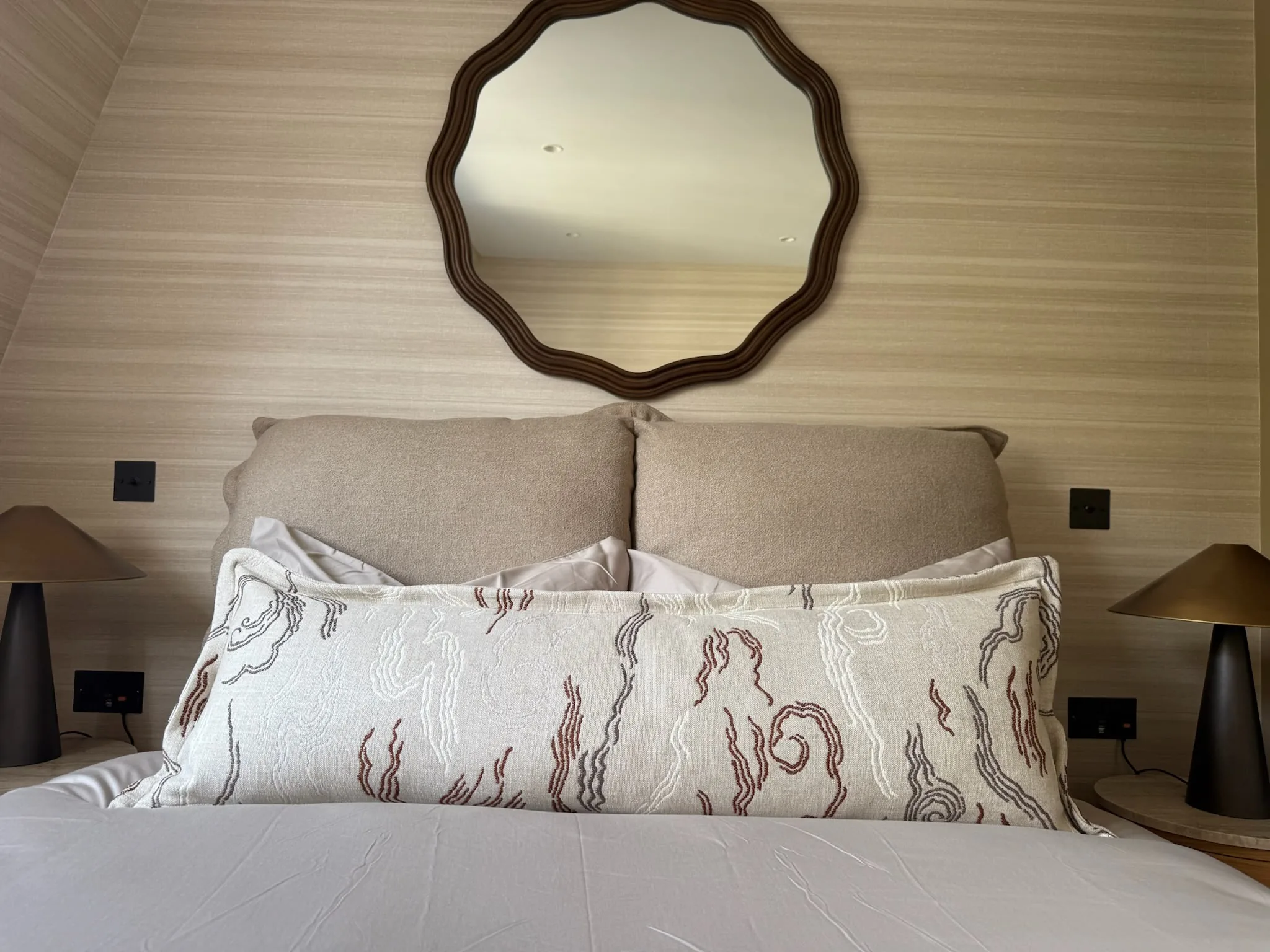 Bed with a padded neutral headboard and a long patterned bolster, under a scalloped wooden mirror