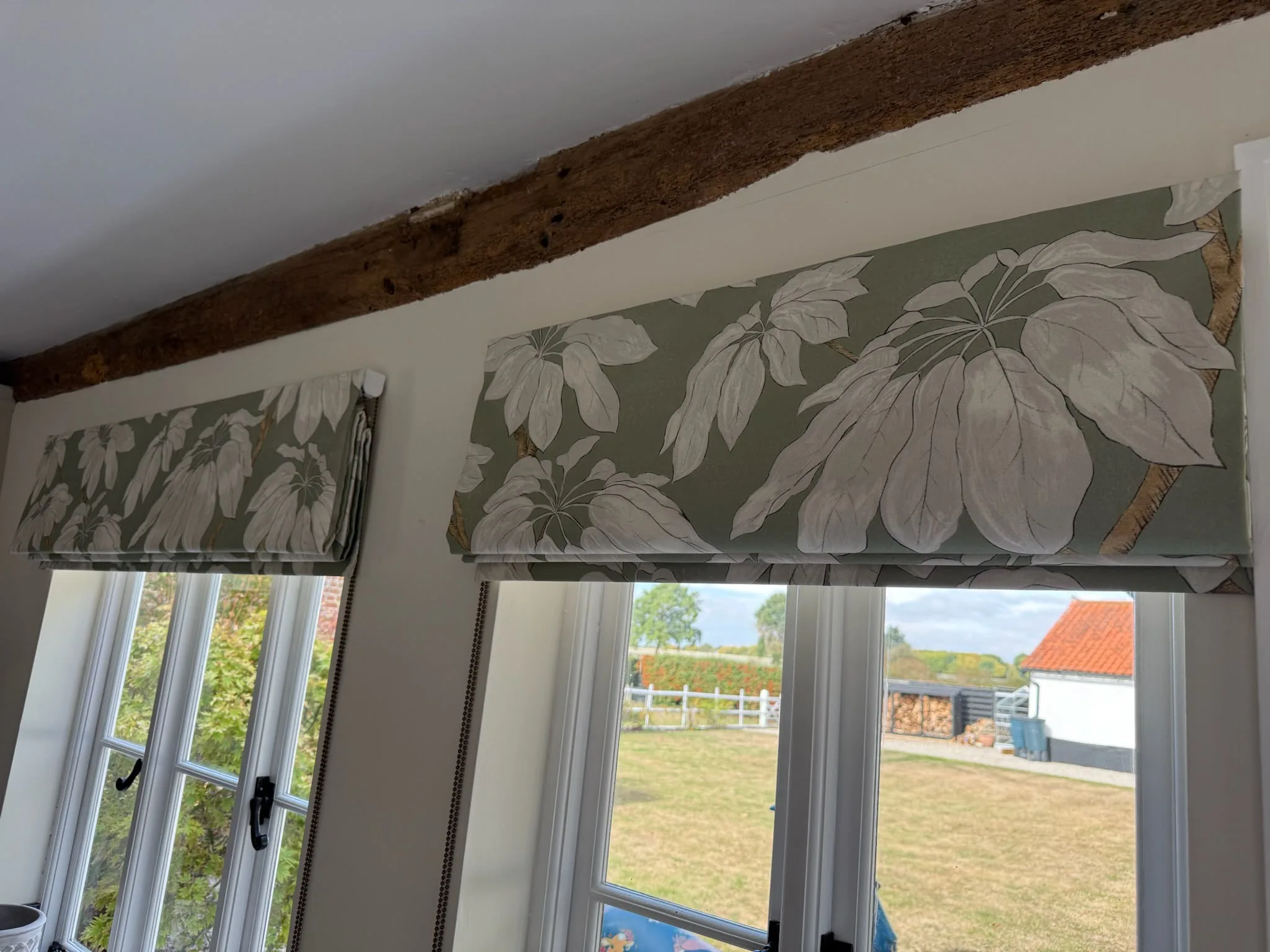 Two Roman blinds in a large sage leaf print beneath an exposed oak beam over casement windows