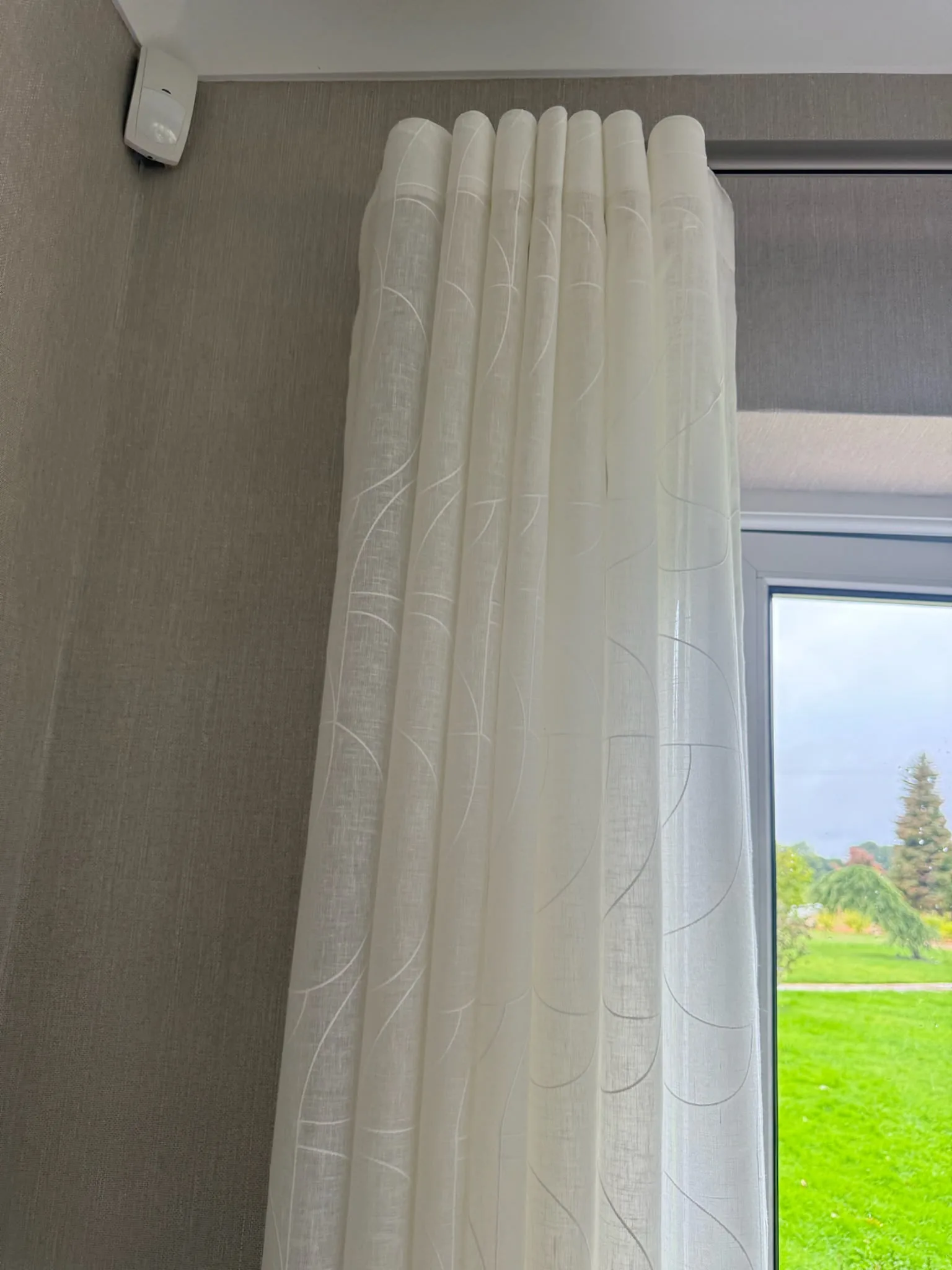 Patterned white voile curtain hanging in tall folds beside a window onto a lawn