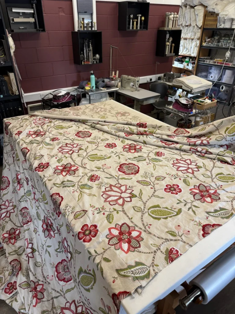 Floral crewel-style fabric laid out on the workroom table, with shelves of tools and supplies behind