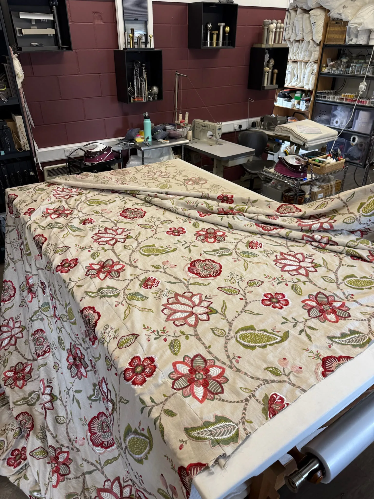 Floral crewel-style fabric laid out on the workroom table, with shelves of tools and supplies behind