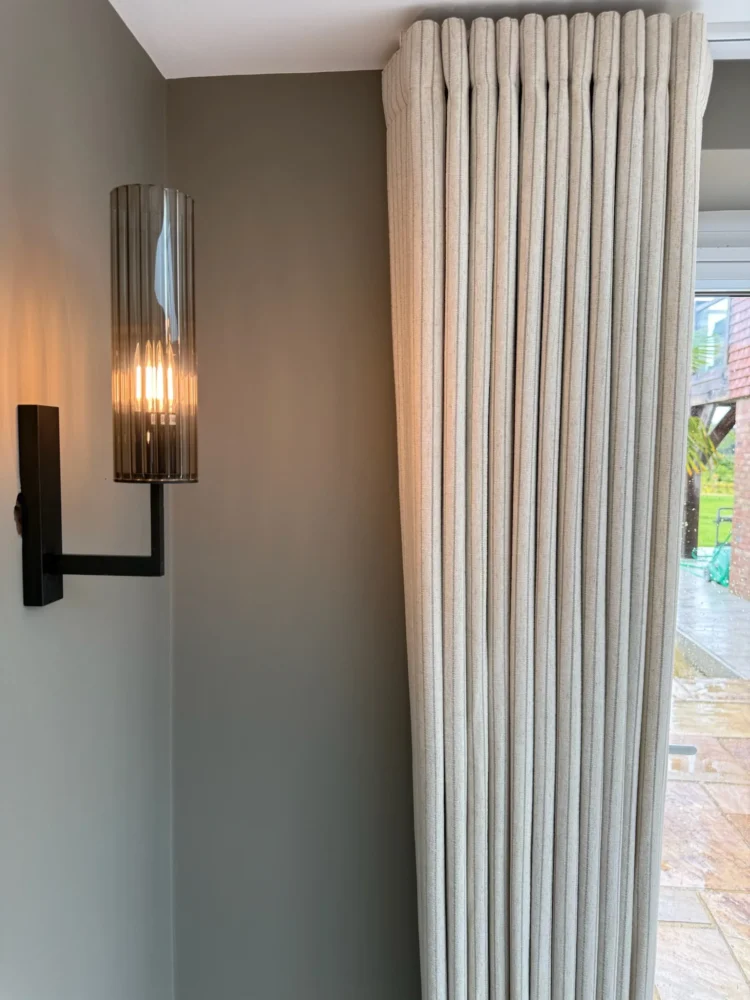 Tall neutral curtain with closely spaced rounded pleats, stacked beside a smoked-glass wall light