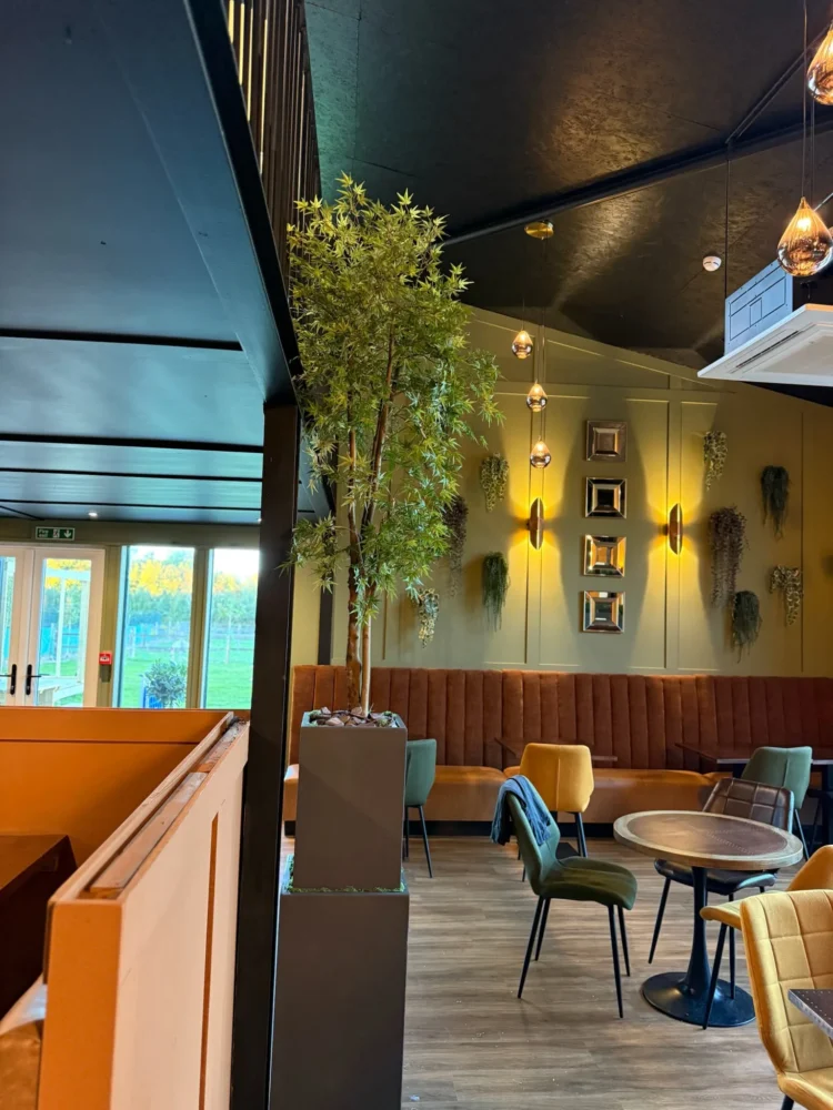 Restaurant with channel-stitched banquette seating, small round tables and a tall indoor tree under a dark ceiling