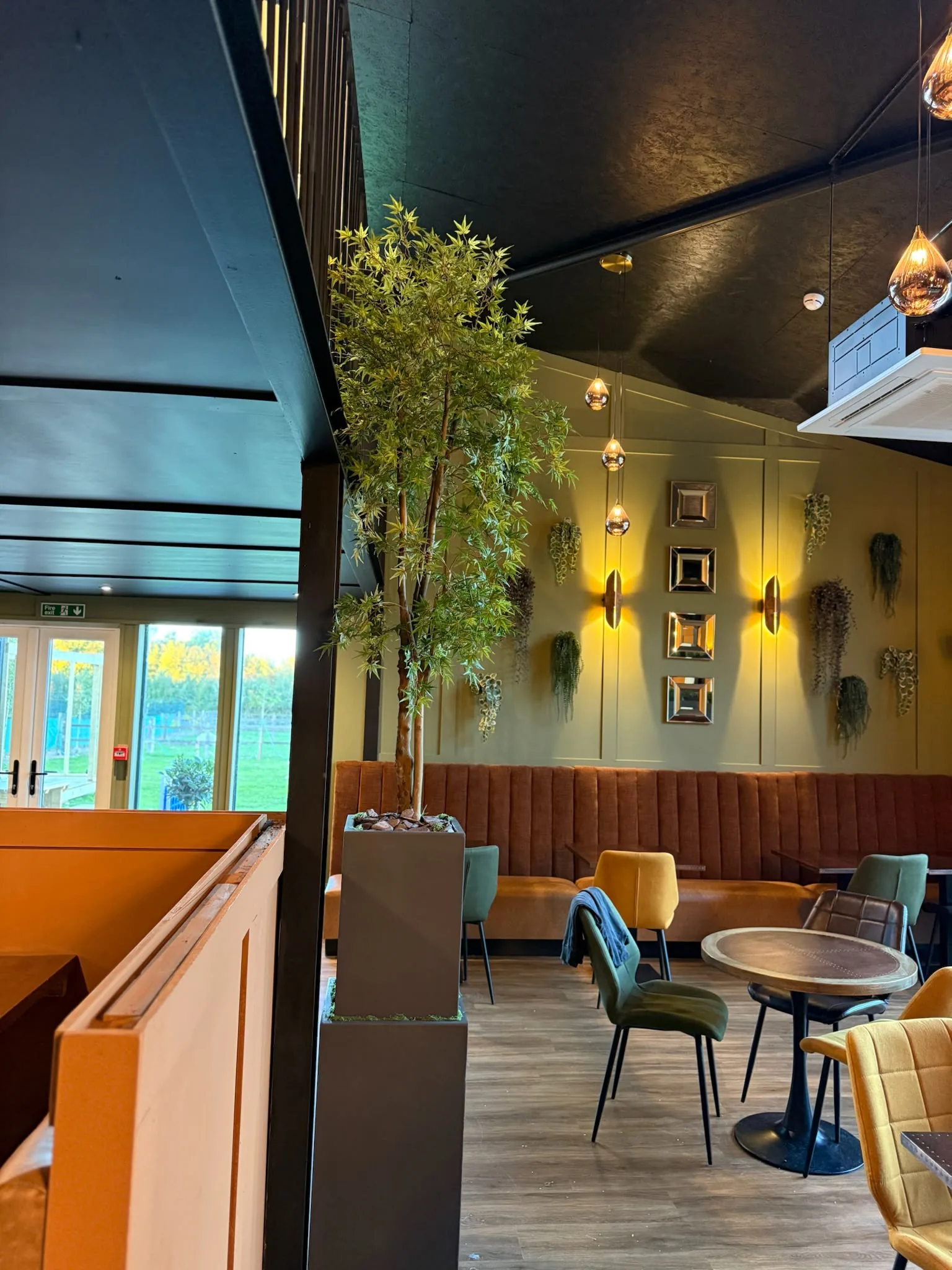 Restaurant with channel-stitched banquette seating, small round tables and a tall indoor tree under a dark ceiling