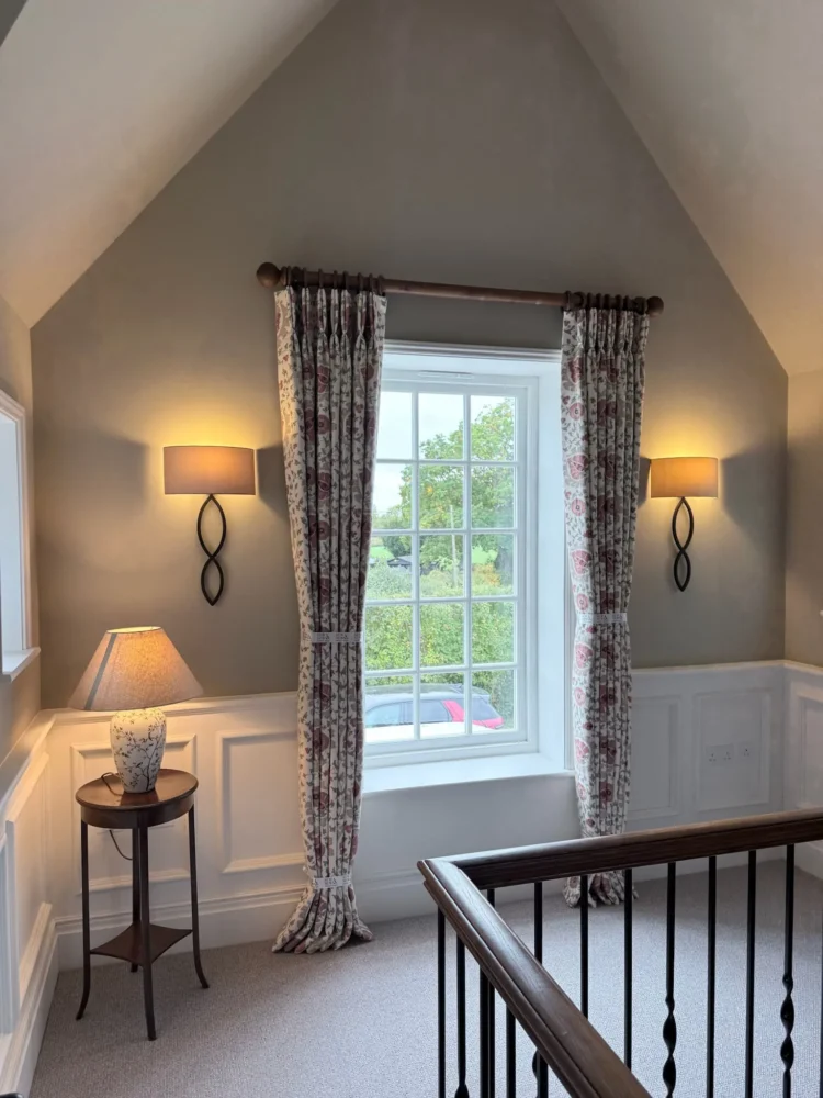 Floral pinch-pleat curtains with tiebacks on a wooden pole at a landing window, between two wall lights