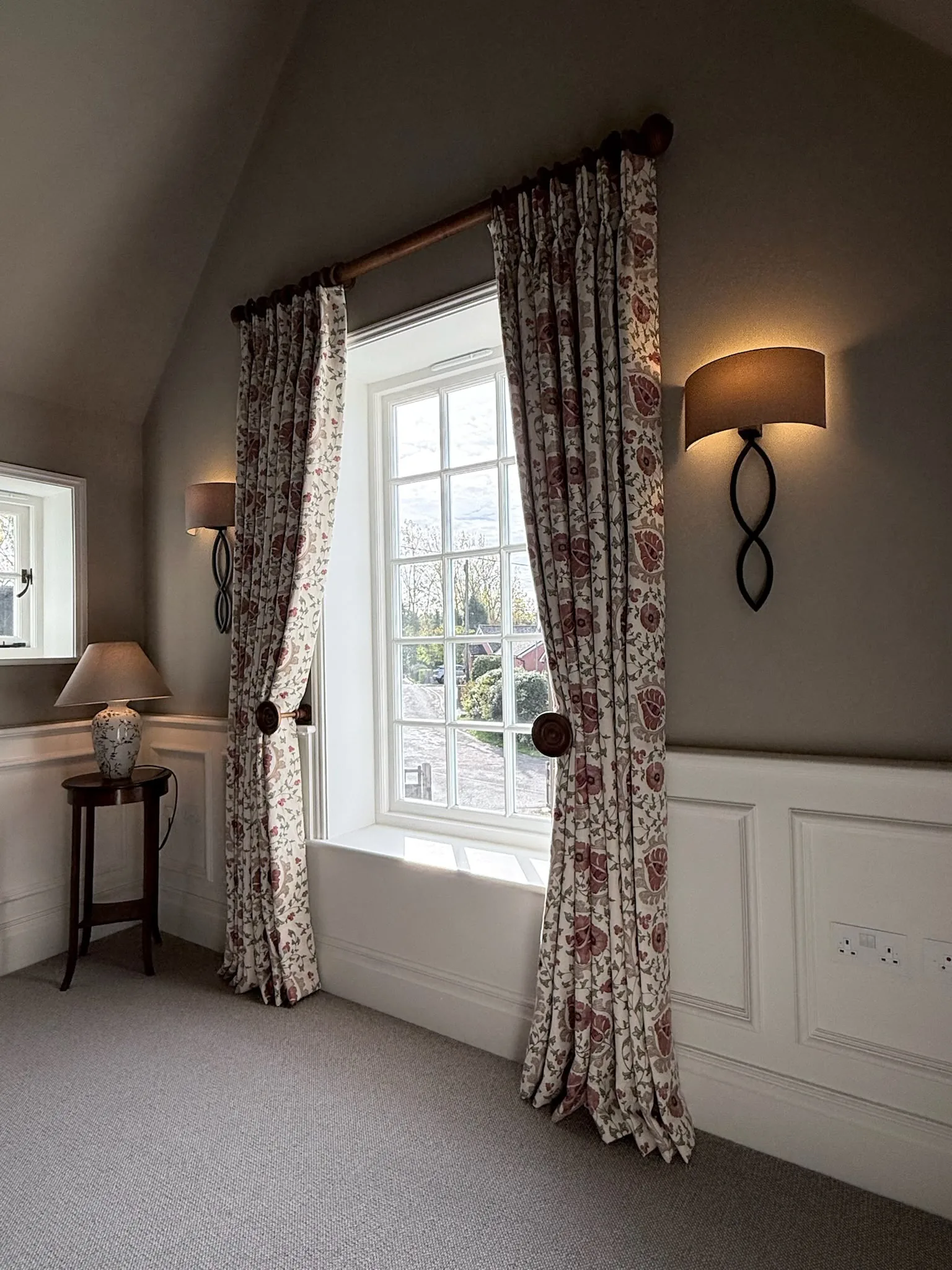 Floral curtains held back with a tieback at a landing window, beside a wall light and side table