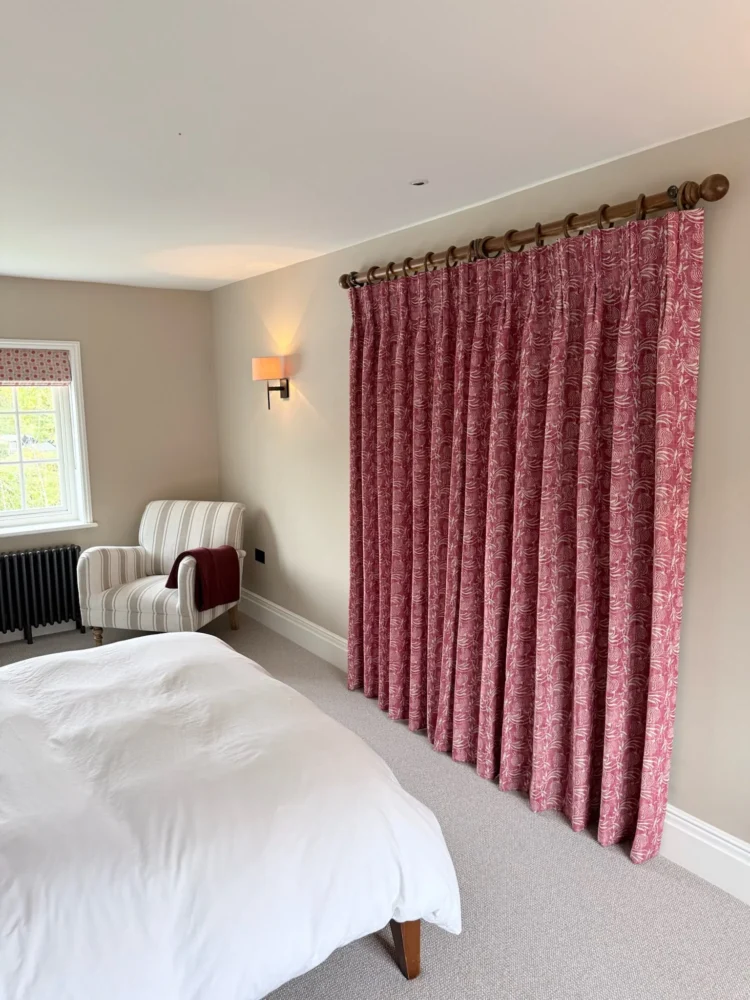 Red patterned pinch-pleat curtains on a wooden pole in a bedroom with a striped armchair