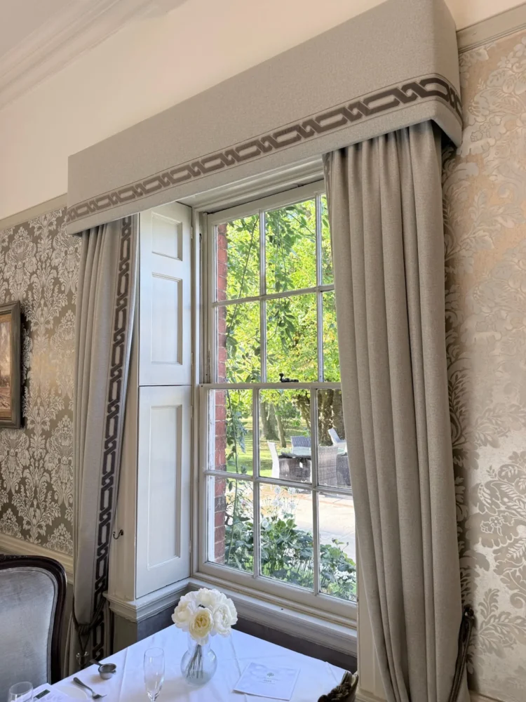 Upholstered pelmet with a Greek key braid over full-length neutral curtains at a sash window, in a dining room with damask wallpaper