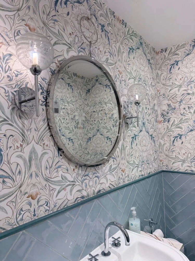 Bathroom with botanical wallpaper, a round chrome mirror between wall lights and sage tiling above a basin