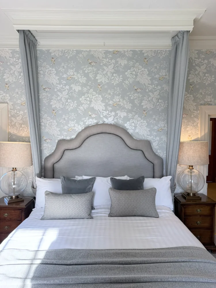 Bed with a shaped upholstered headboard framed by a canopy and curtain drapes, against blue floral wallpaper