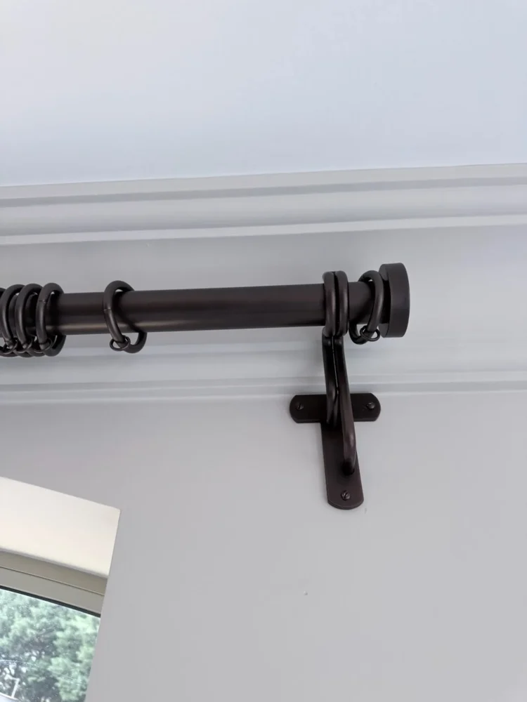 Close-up of a dark bronze curtain pole with rings and a passing bracket, fixed below a cornice