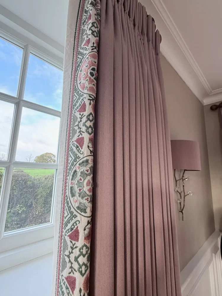 Dusky pink pinch-pleat curtain with a printed floral border, hanging beside a sash window