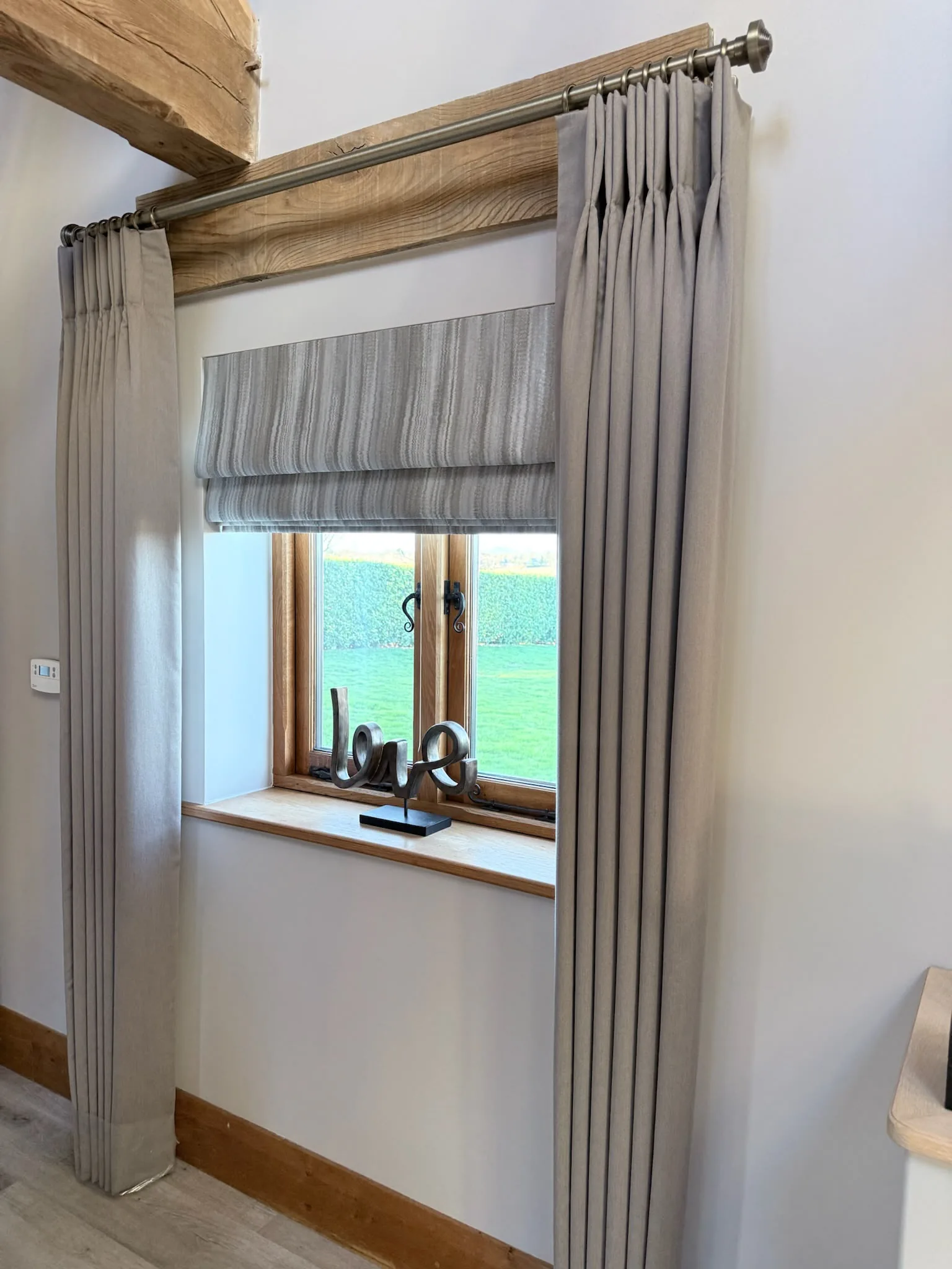 Grey pinch-pleat dress curtains on a metal pole framing a striped Roman blind in an oak-framed window