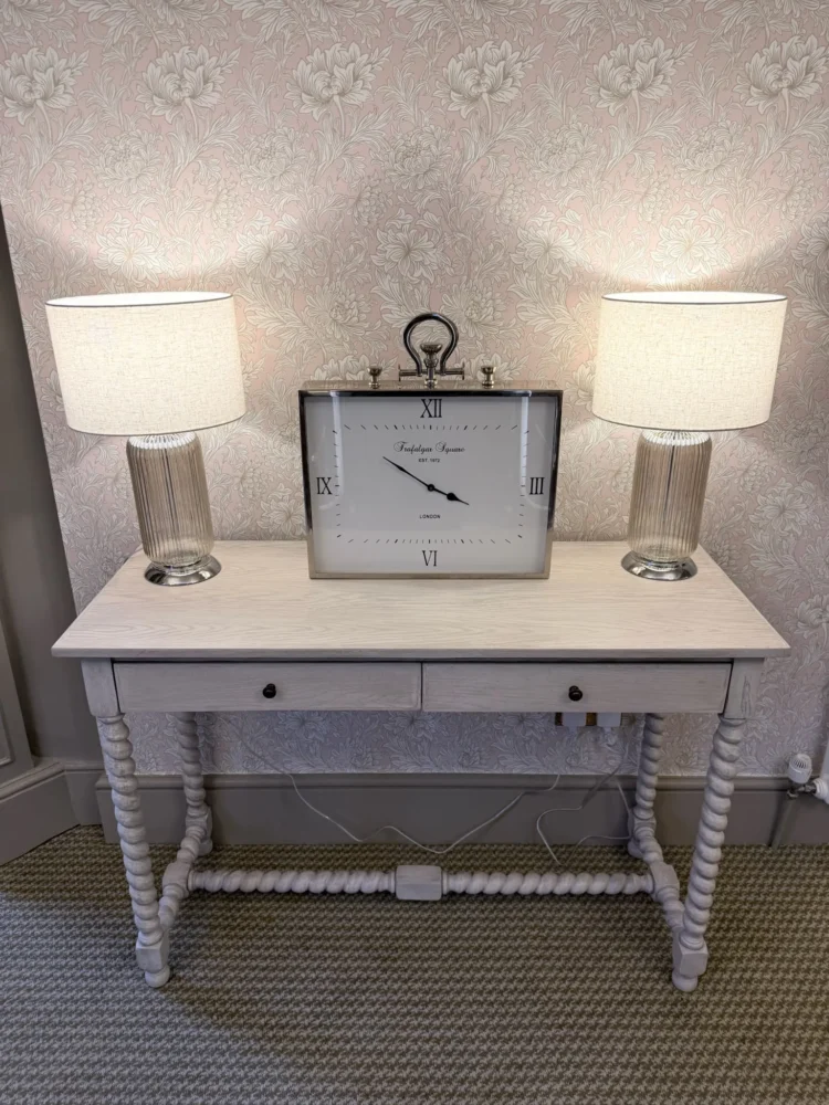 Painted console table with two glass lamps and a large clock against pink floral wallpaper