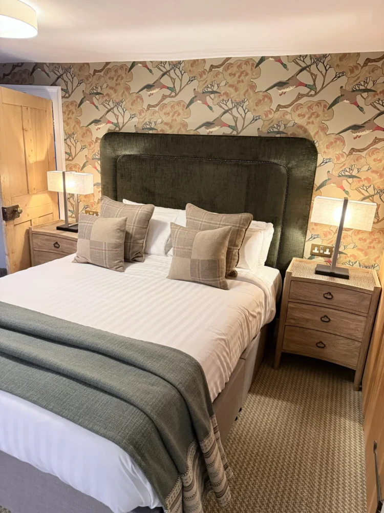 Bed with a tall green upholstered headboard, check cushions and a teal throw against bird and blossom wallpaper