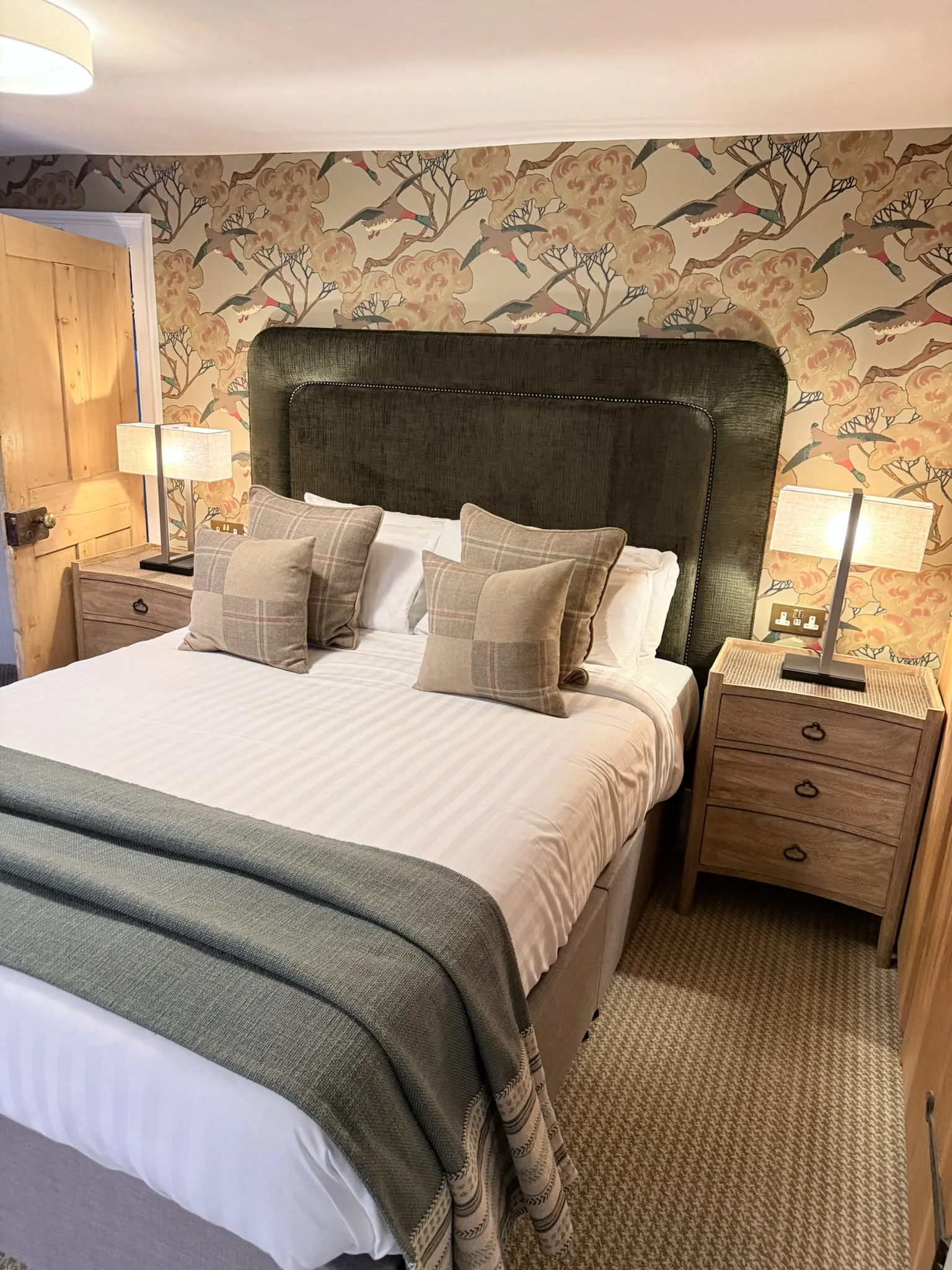 Bed with a tall green upholstered headboard, check cushions and a teal throw against bird and blossom wallpaper