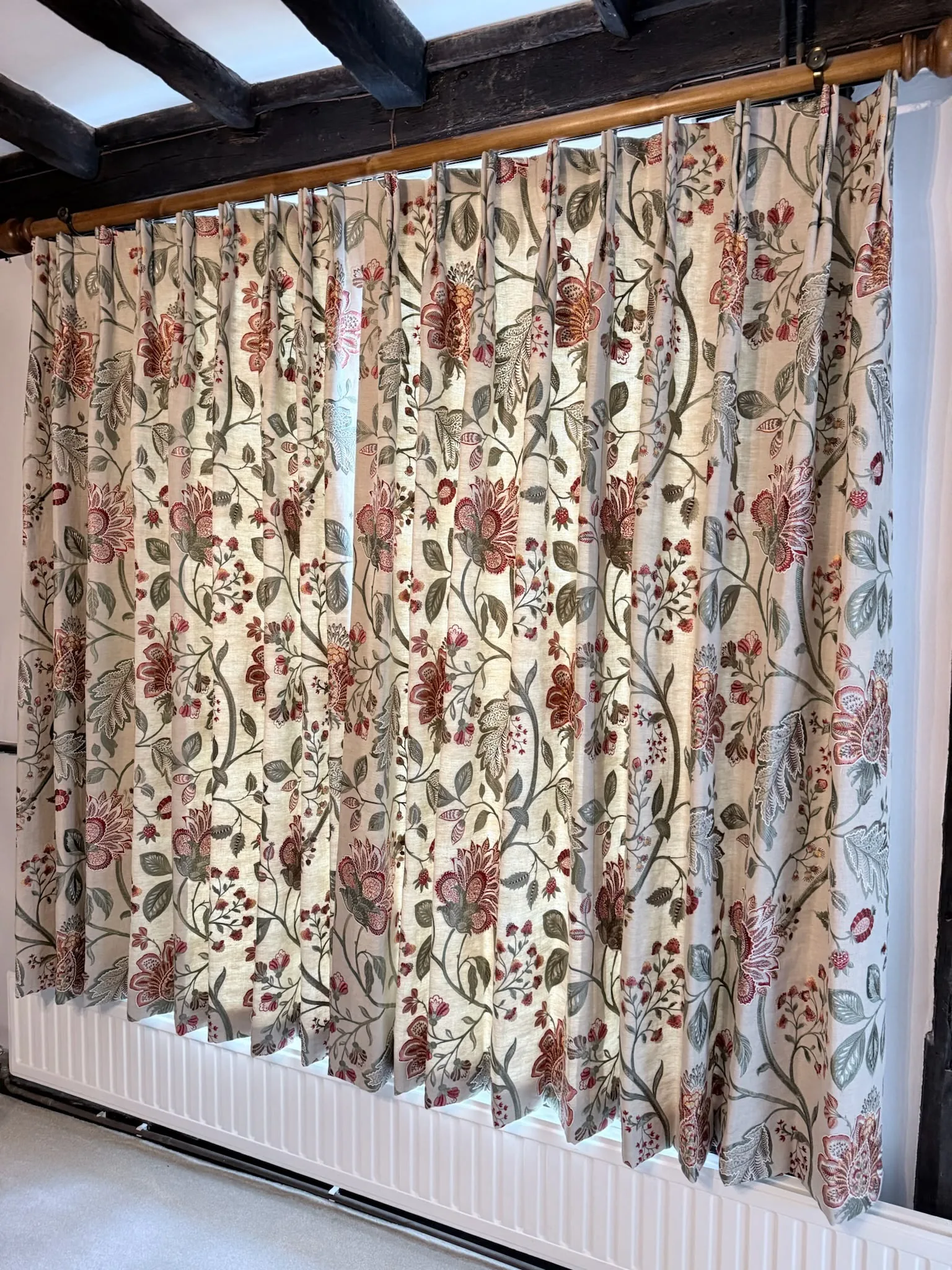 Floral crewel-style pinch-pleat curtains drawn across a window under exposed beams