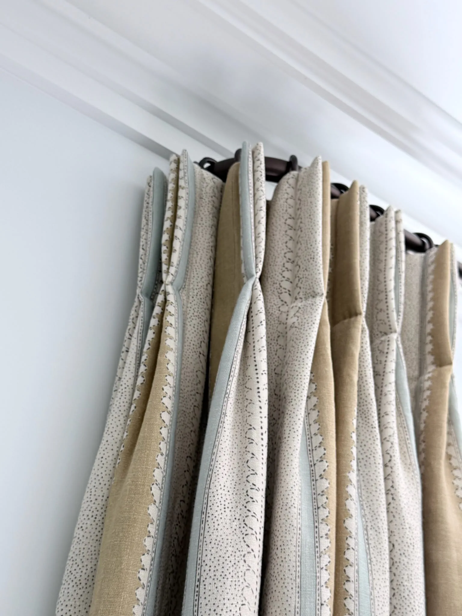 Pinch-pleat curtains in a neutral striped and spotted fabric, on rings on a dark pole below a cornice