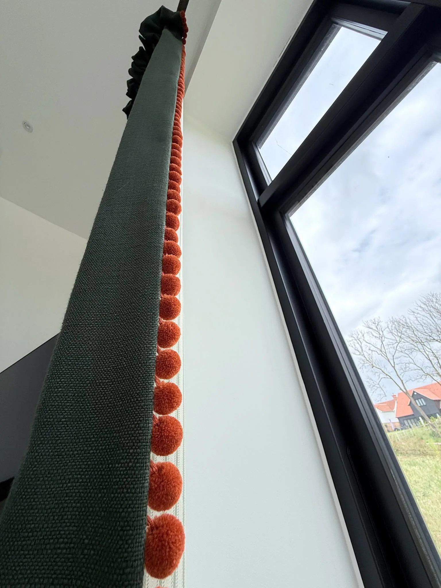 Looking up a dark green curtain edged with orange pom-poms beside tall black-framed windows