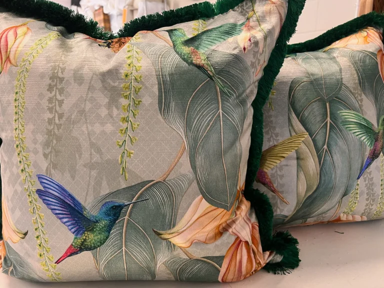 Pair of cushions in a hummingbird and tropical leaf print, edged with green fringe