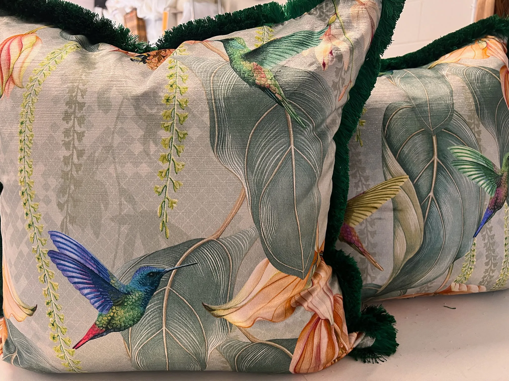 Pair of cushions in a hummingbird and tropical leaf print, edged with green fringe