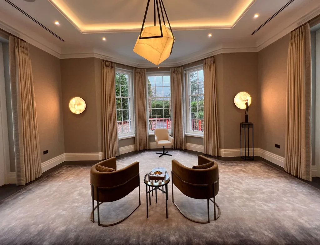 Large reception room with floor-length neutral curtains at every window, two tan tub chairs, a statement pendant and alabaster wall lights