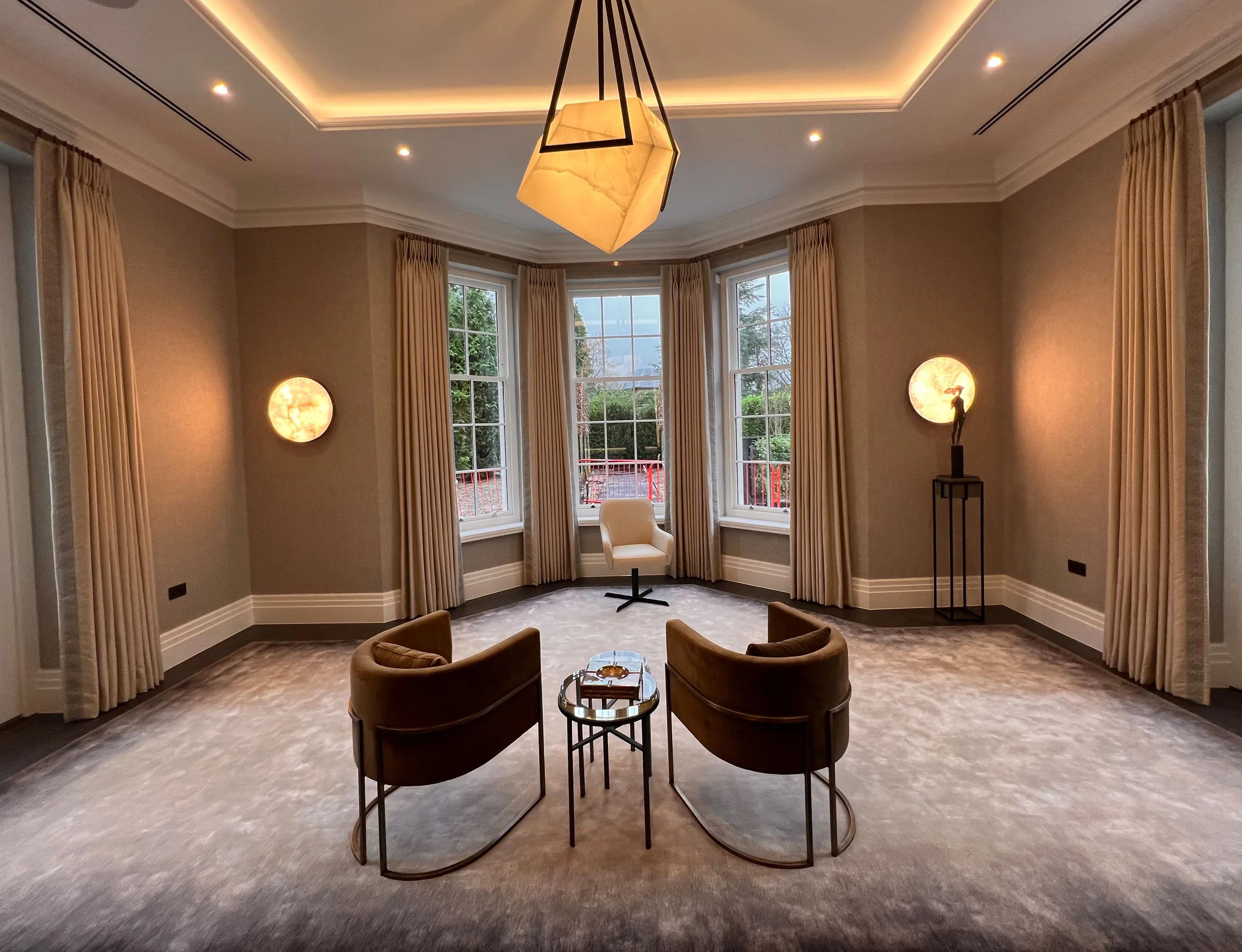 Large reception room with floor-length neutral curtains at every window, two tan tub chairs, a statement pendant and alabaster wall lights