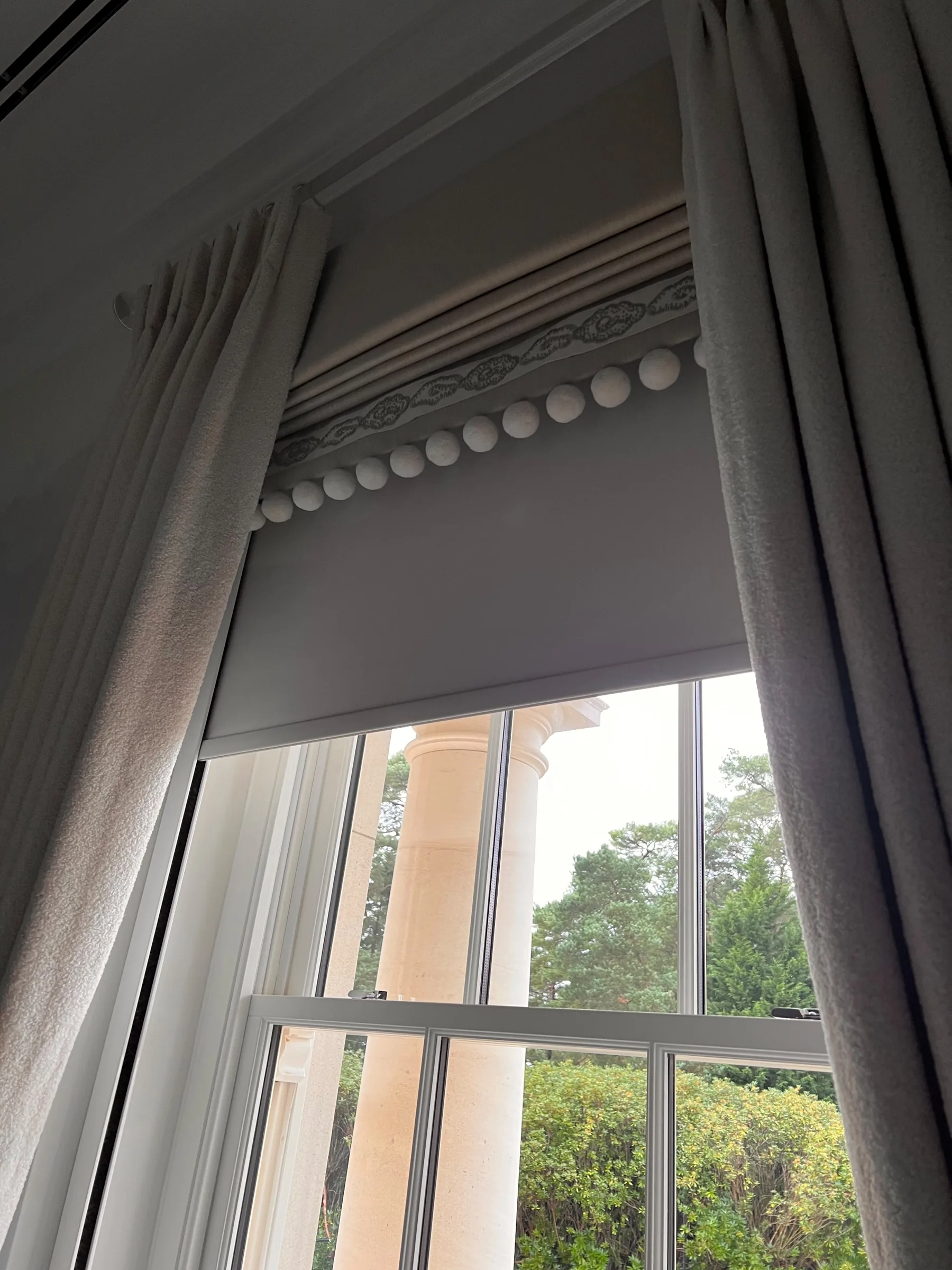 Grey Roman blind with a bobble trim between grey dress curtains at a sash window