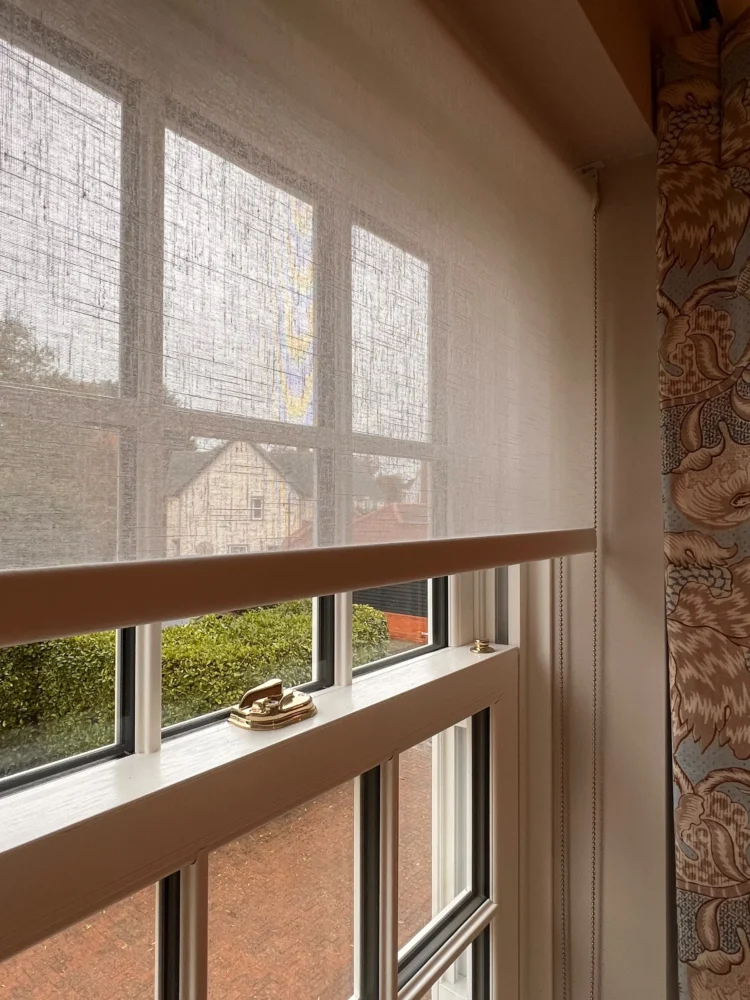 Sheer linen-look roller blind half lowered over a sash window, beside a patterned curtain