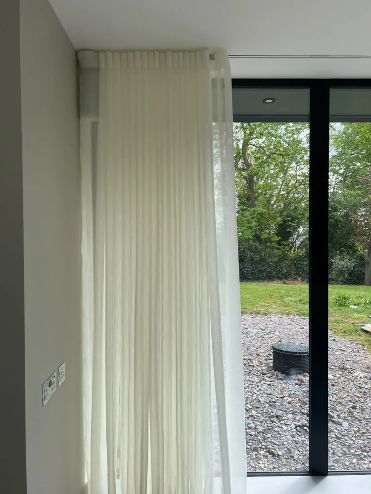 Floor-length sheer voile on a ceiling track beside black-framed glazing onto a garden