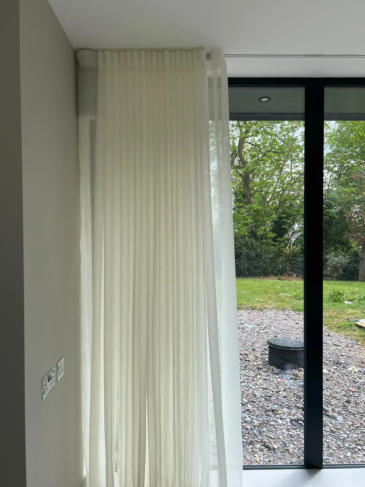 Floor-length sheer voile on a ceiling track beside black-framed glazing onto a garden
