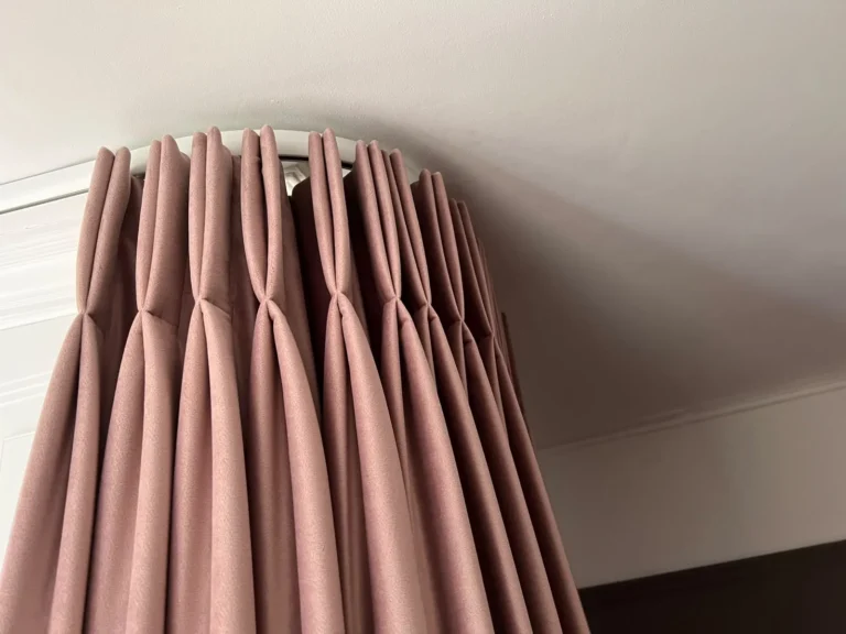 Dusky pink pinch-pleat curtains seen from below on a curved track beneath a cornice