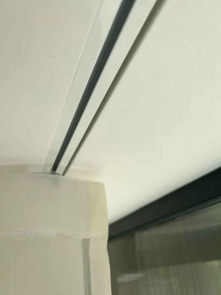 Close-up of a slim ceiling-mounted track carrying a sheer voile heading, beside black-framed glazing