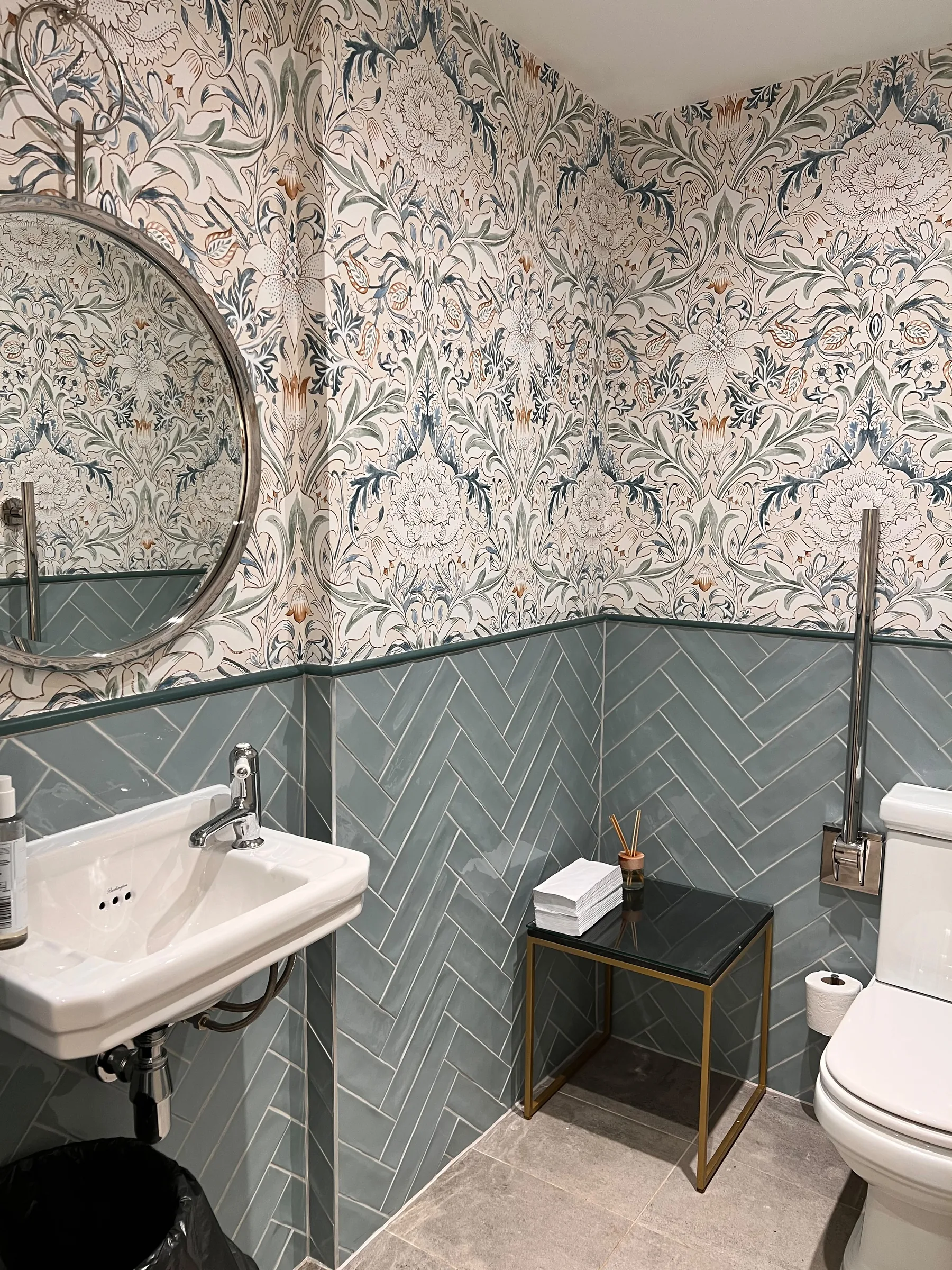 Cloakroom with botanical wallpaper above sage herringbone tiles, a round mirror and a small basin