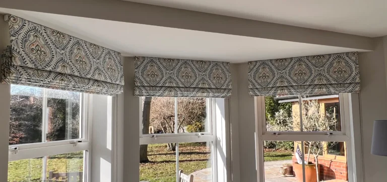 Three Roman blinds in a blue and cream ogee print raised across a square bay of sash windows