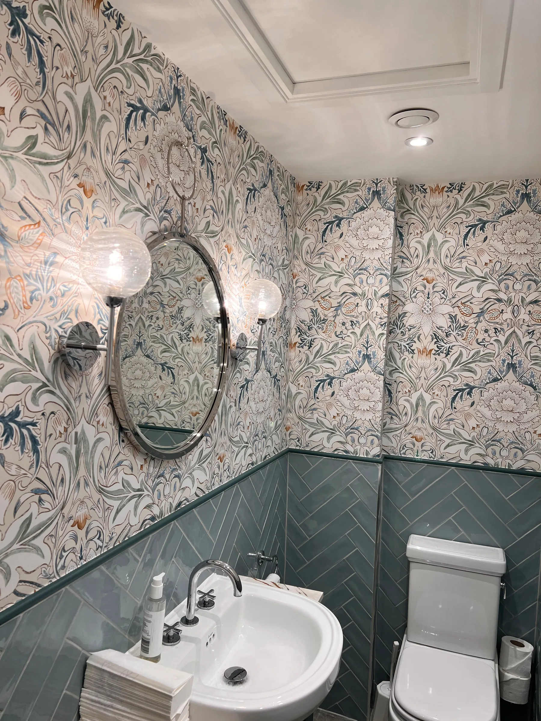 Cloakroom with botanical wallpaper, a round mirror between glass wall lights, a basin and WC