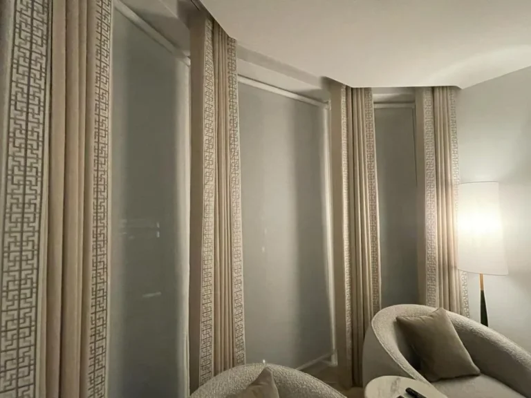 Neutral curtains with Greek key borders around a curved window wall, beside a tub chair
