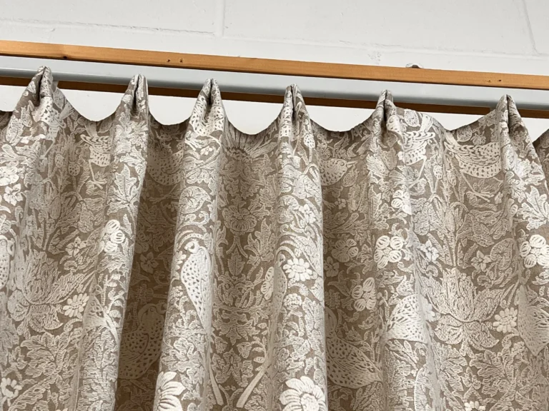 Patterned curtain in a cream and taupe bird and flower design, hung from a batten against a brick wall for checking
