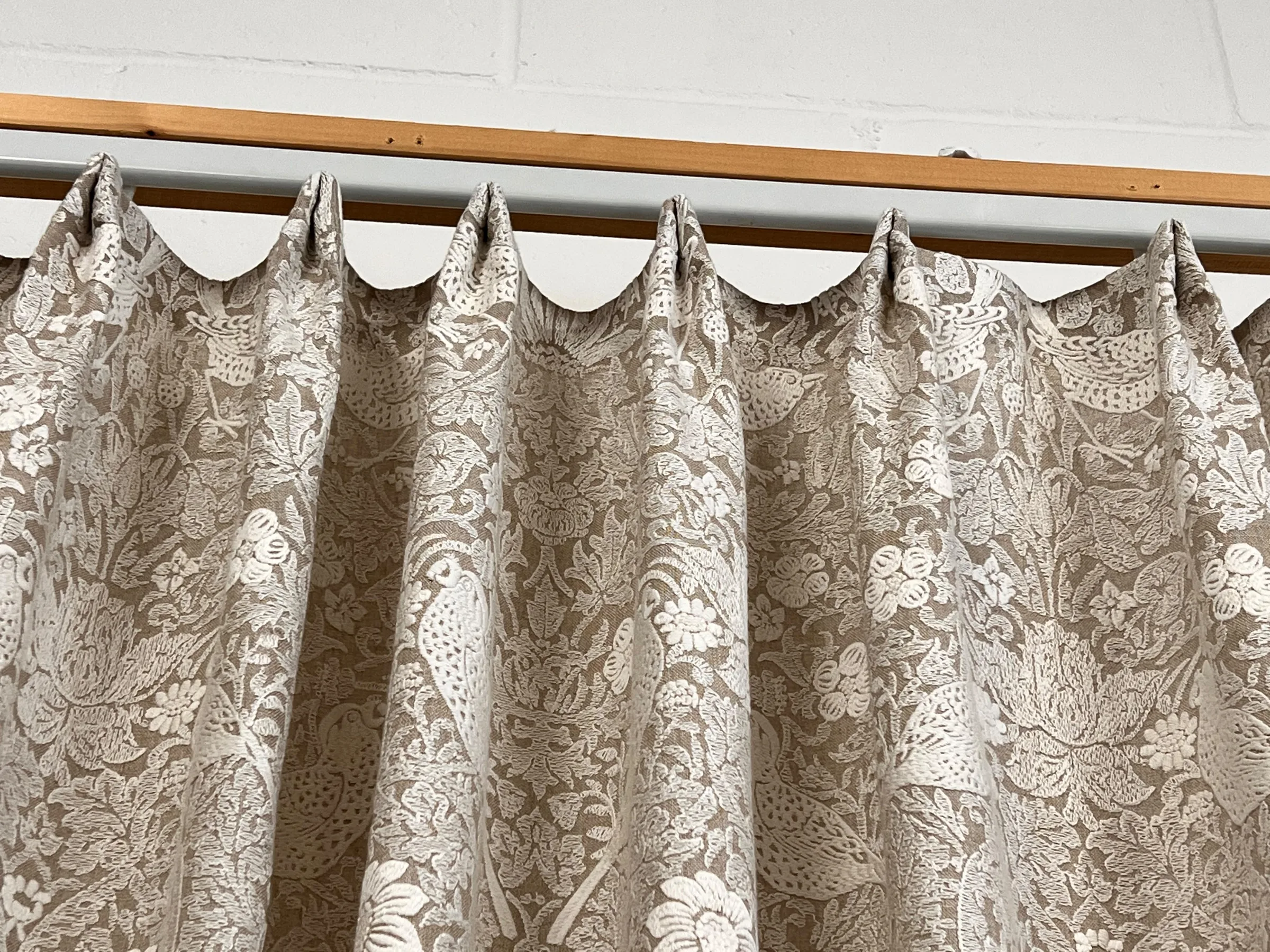 Patterned curtain in a cream and taupe bird and flower design, hung from a batten against a brick wall for checking