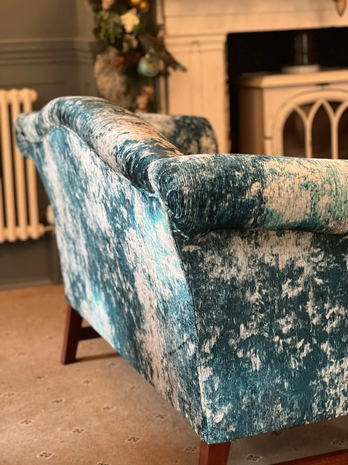 Arm of a sofa upholstered in teal crushed velvet, beside a fireplace and radiator