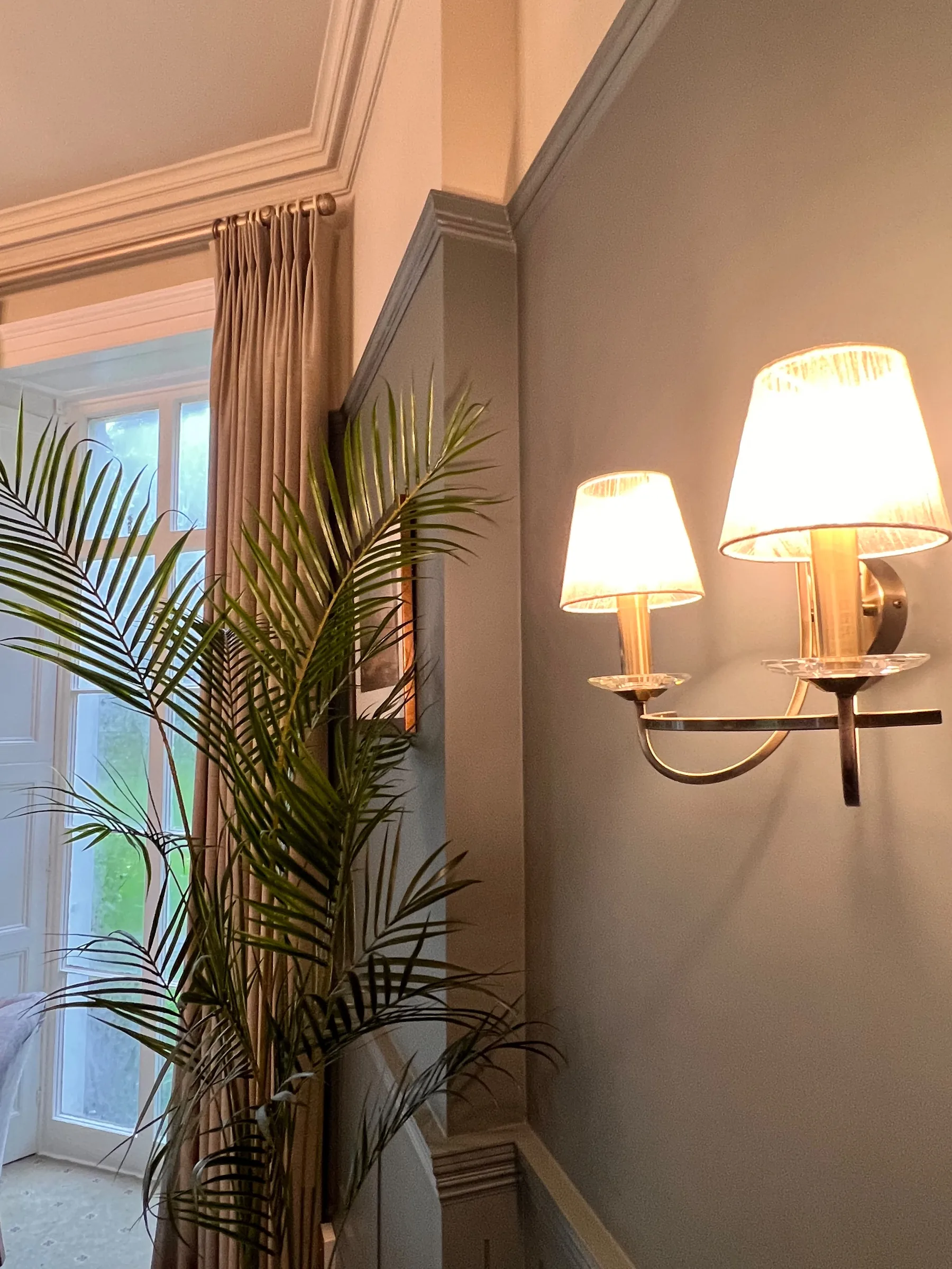 Neutral pinch-pleat curtain on a pole beside a twin-arm wall light and a palm, at Mulberry House