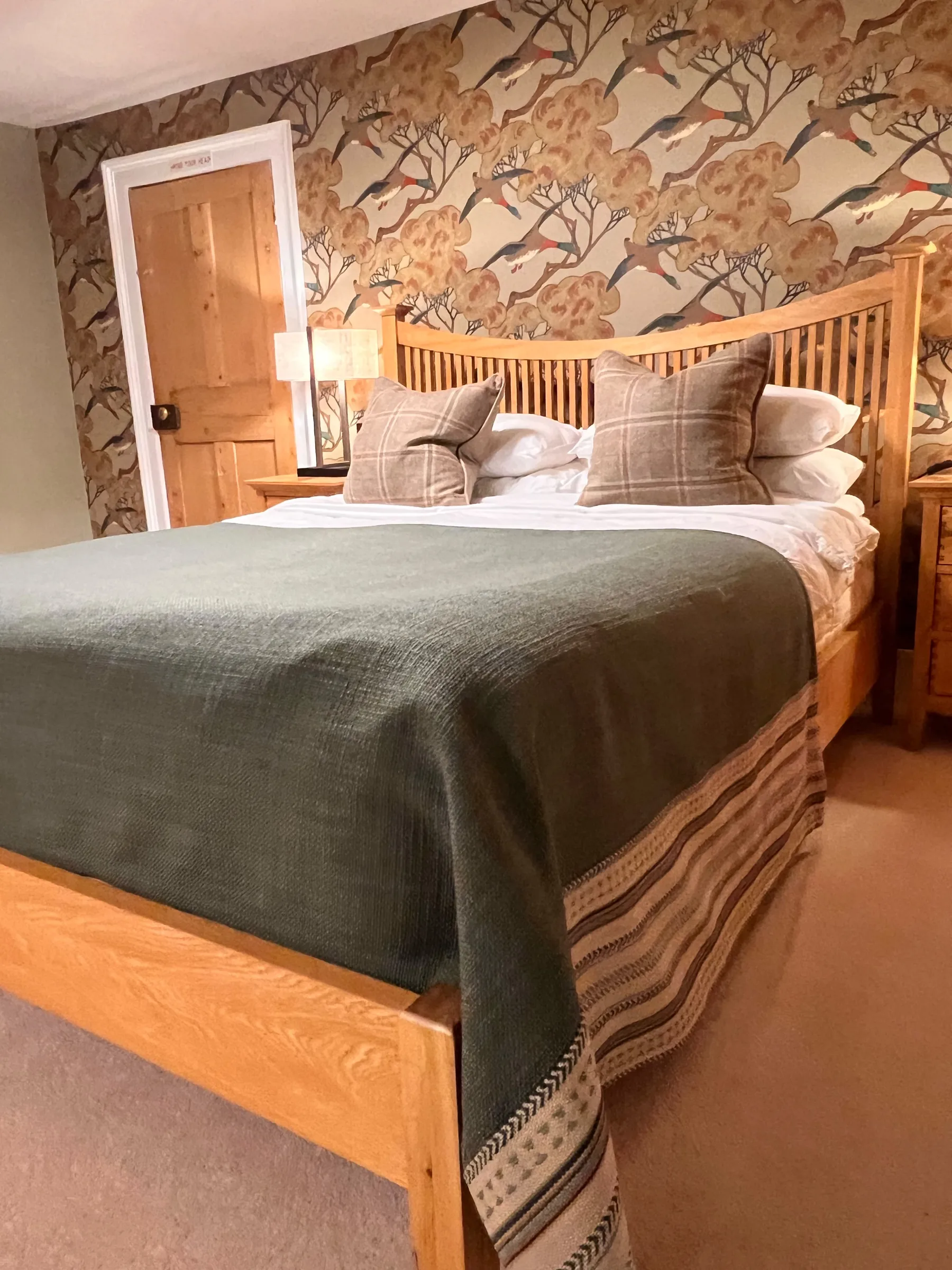 Bed at Mulberry House dressed with a green throw with a striped border and check cushions, against bird and blossom wallpaper