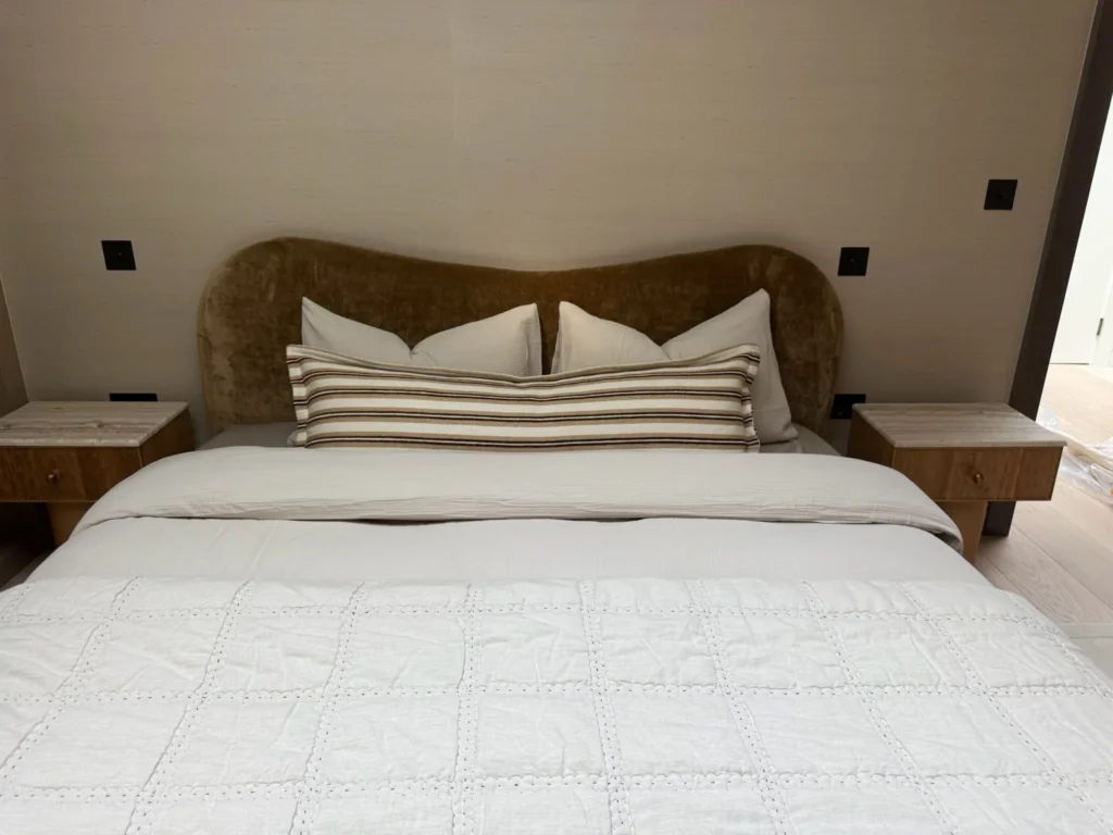 Bed with a curved olive velvet headboard and a striped bolster cushion, between floating bedside tables