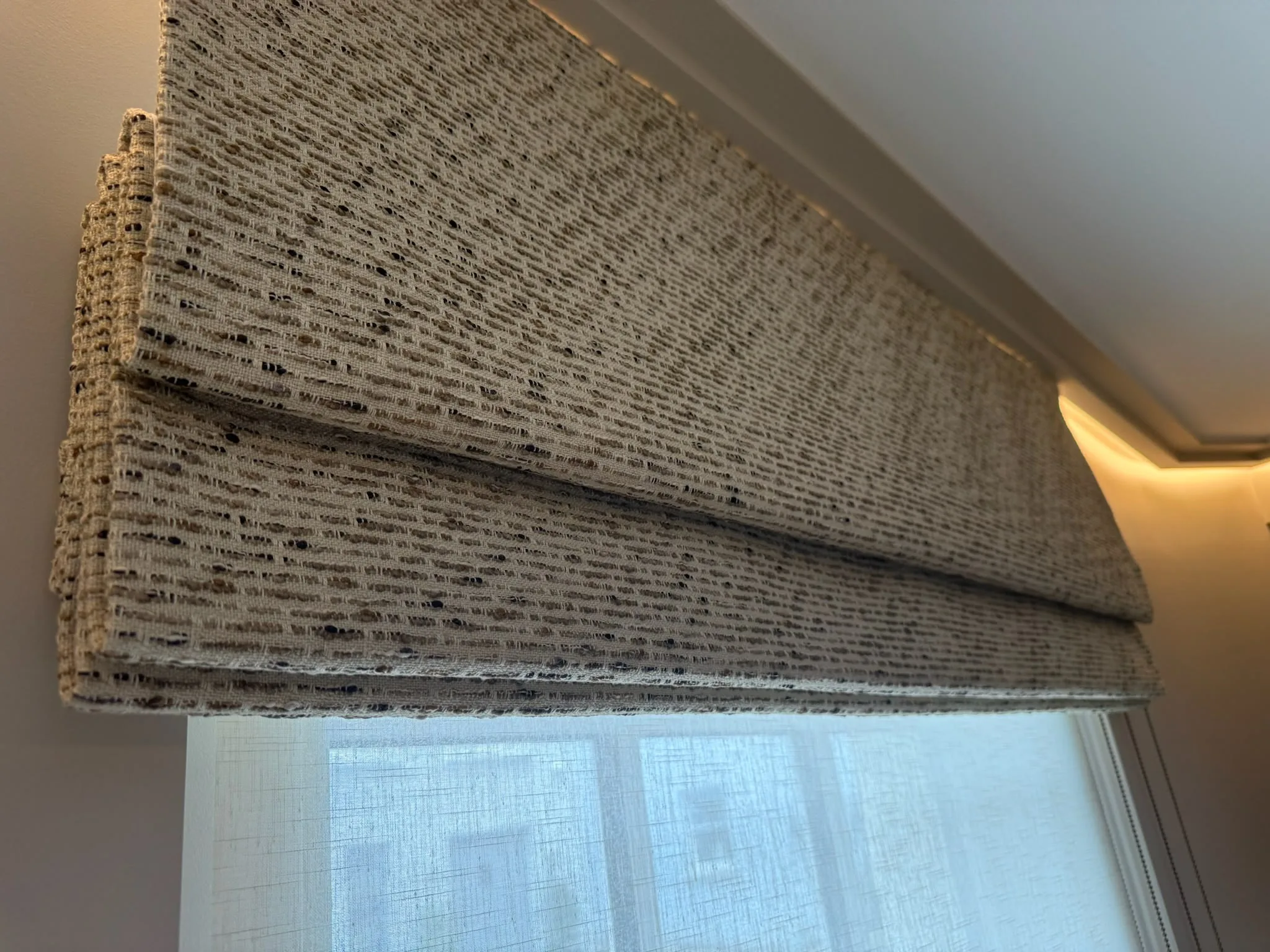Flecked woven Roman blind folded above a window with a sheer blind and concealed lighting