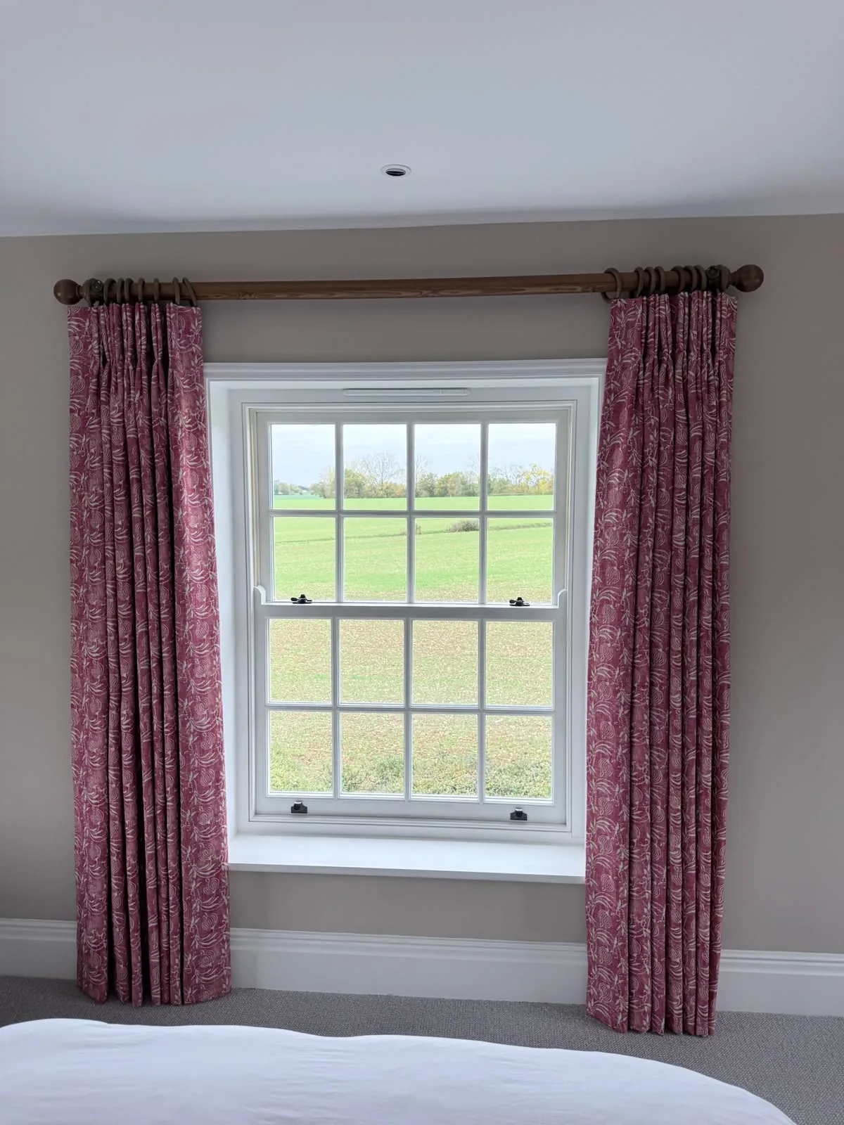 Red patterned pinch-pleat curtains on a wooden pole framing a sash window that looks onto fields