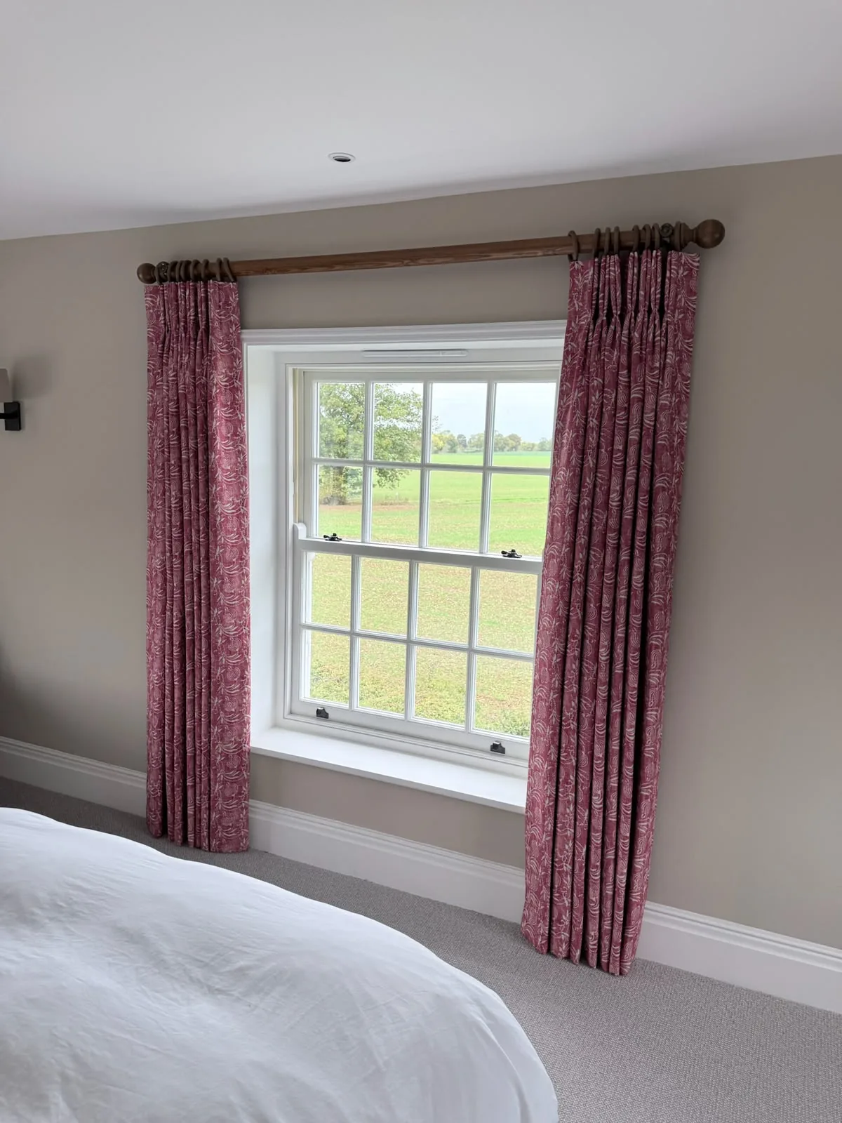 Red patterned curtains open either side of a sash window, seen from beside the bed